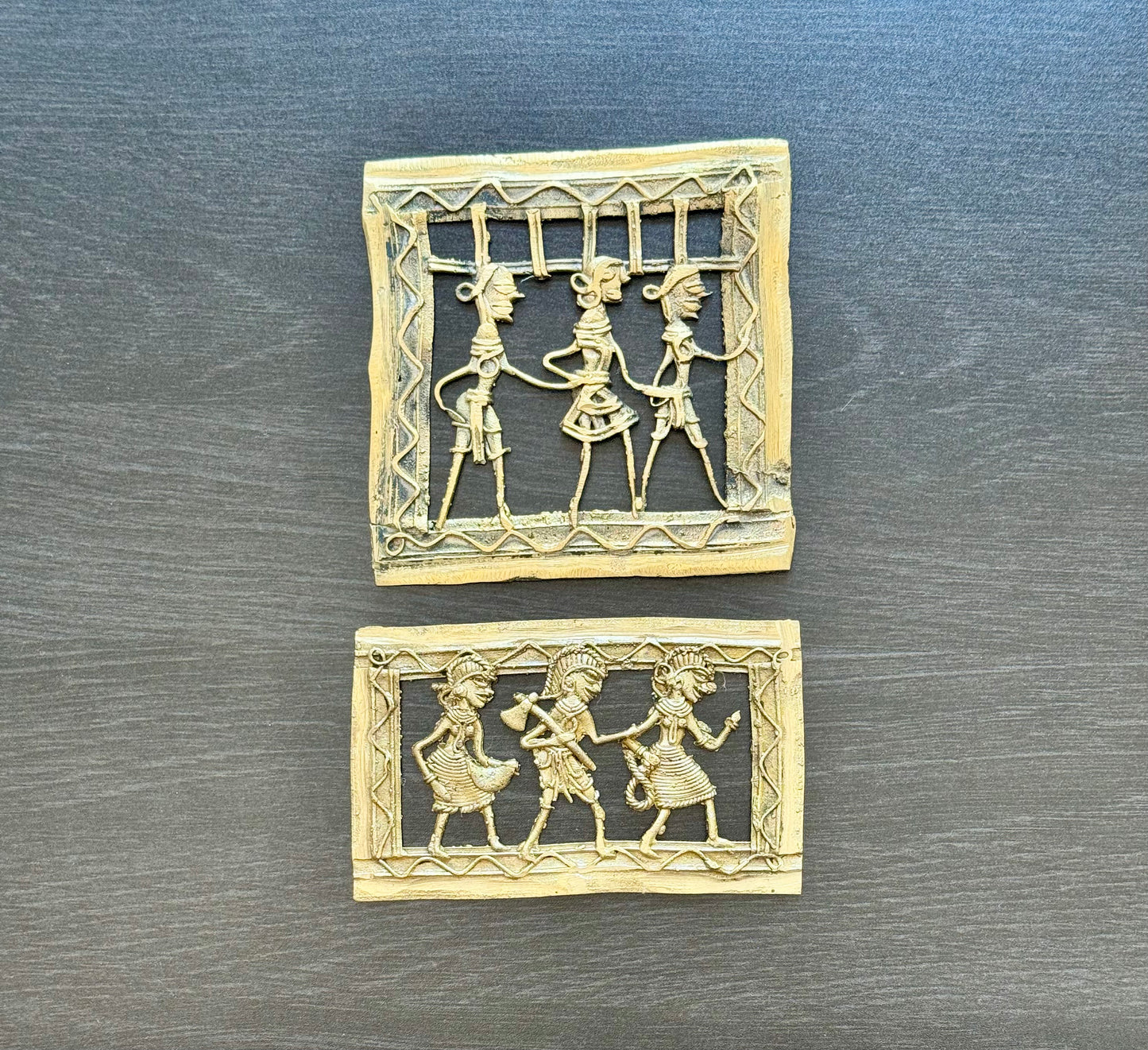 Two small square and rectangular carved brass Dhokra pieces with cutout figures, displayed on a dark gray wood-textured surface.