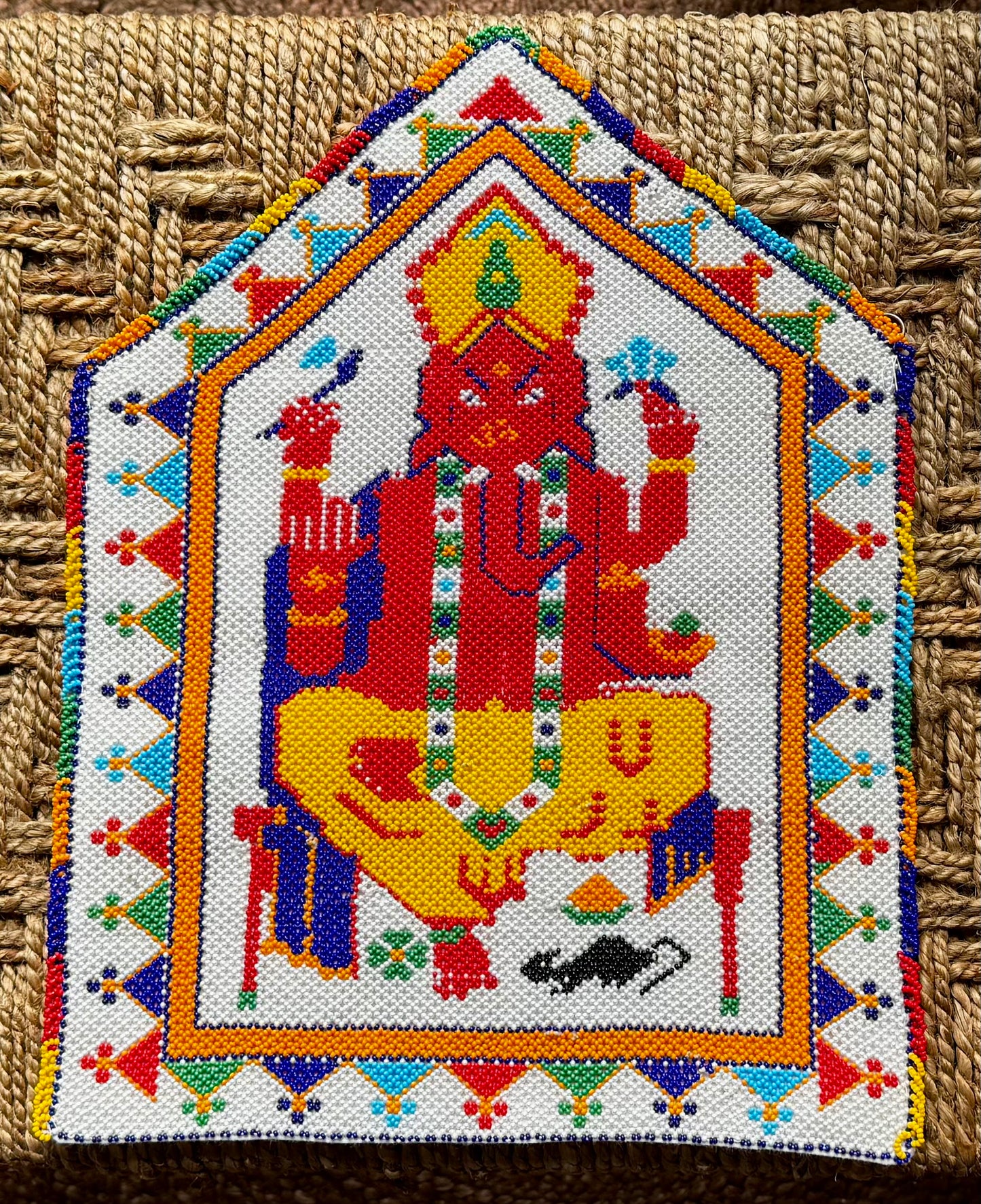 Decorative Motikaam - strung Pearl work Ganesha wall hanging with colorful design on a textured fabric background. Colorful embroidered artwork of a seated figure with geometric patterns on a textured surface
