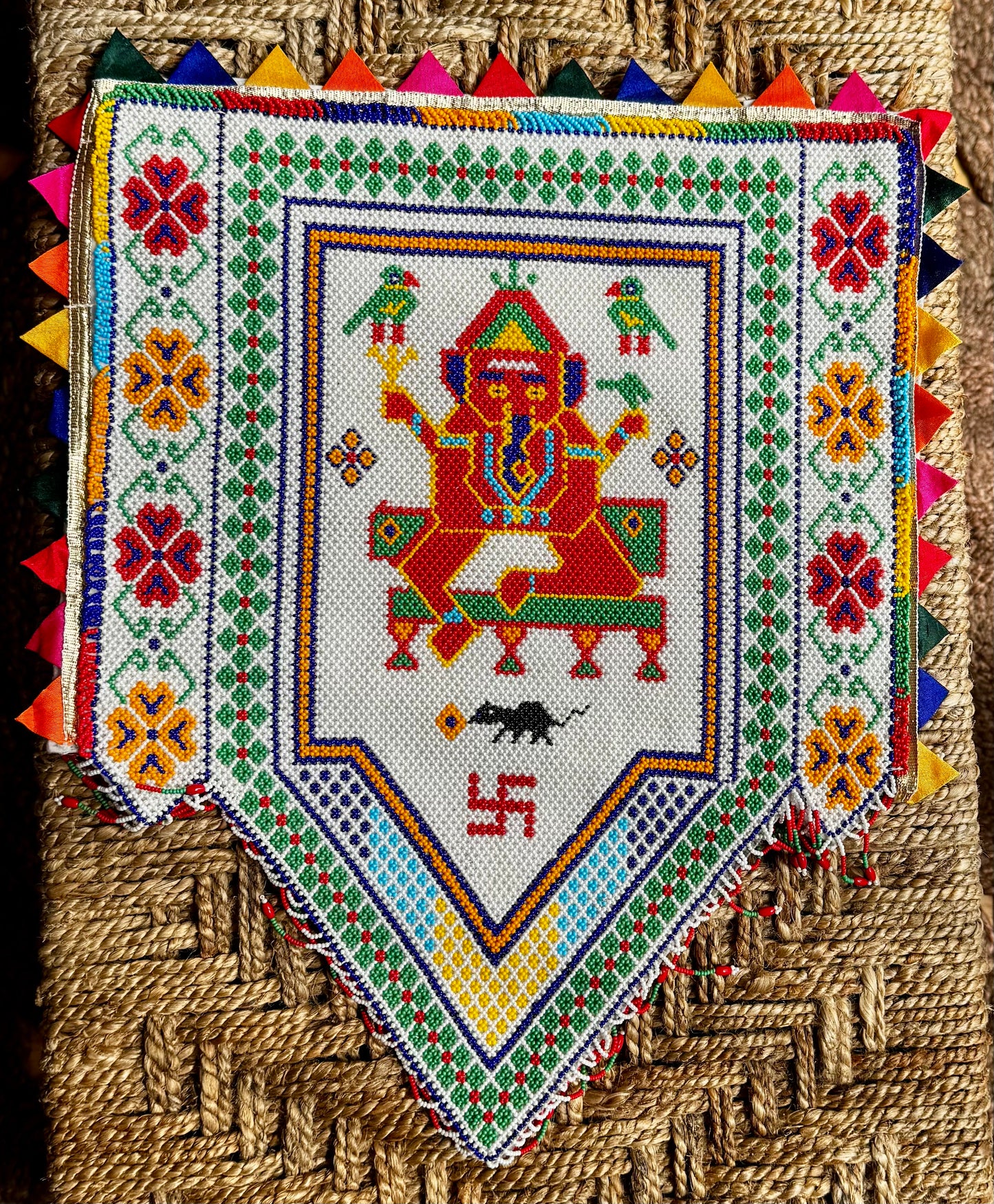 Decorative Motikaam - strung Pearl work Ganesha wall hanging with colorful design on a textured fabric background. Colorful embroidered fabric with a central figure and geometric patterns on a woven background