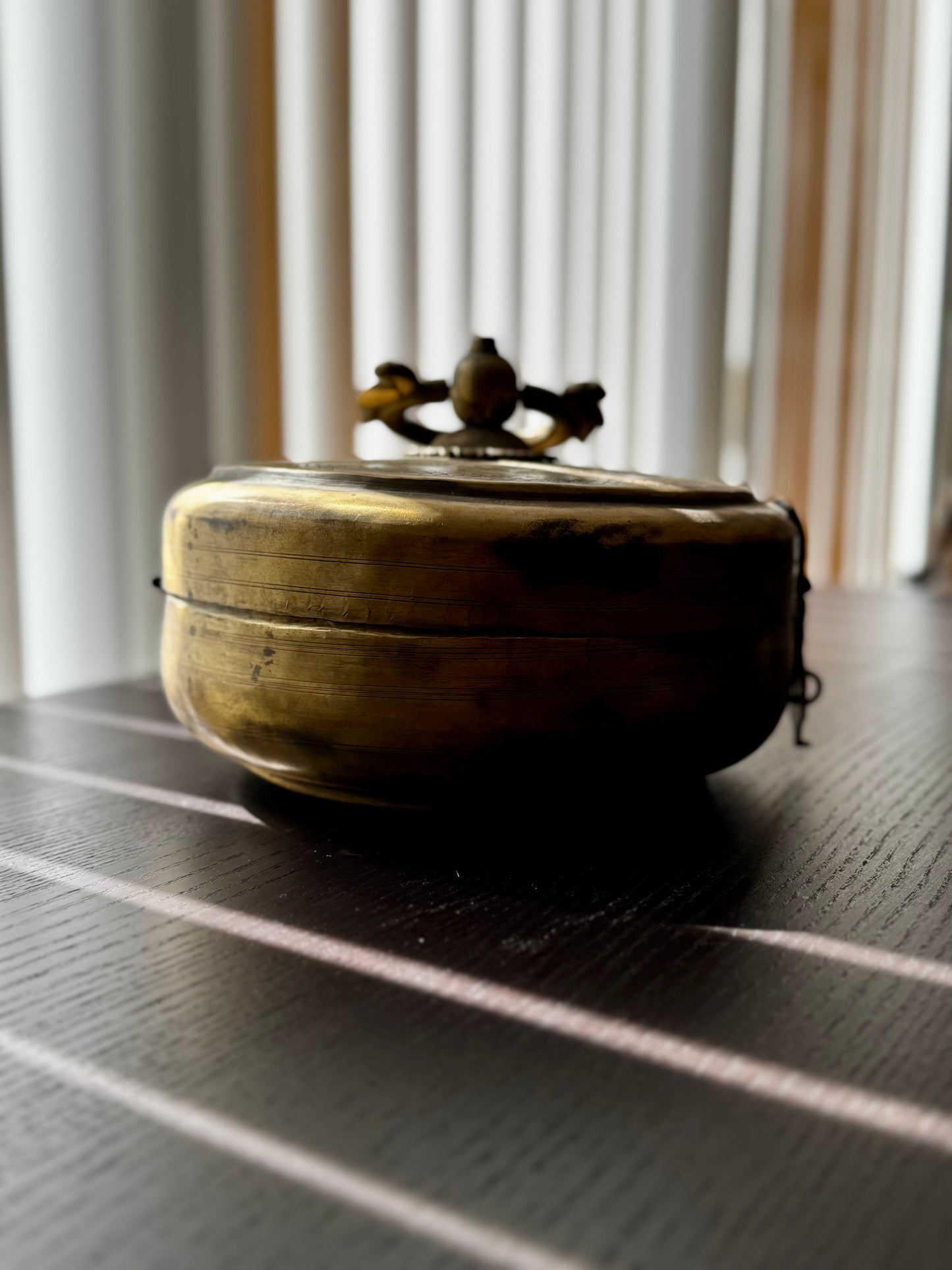 Brass box with lid on a dark surface with light filtering through curtains in the background