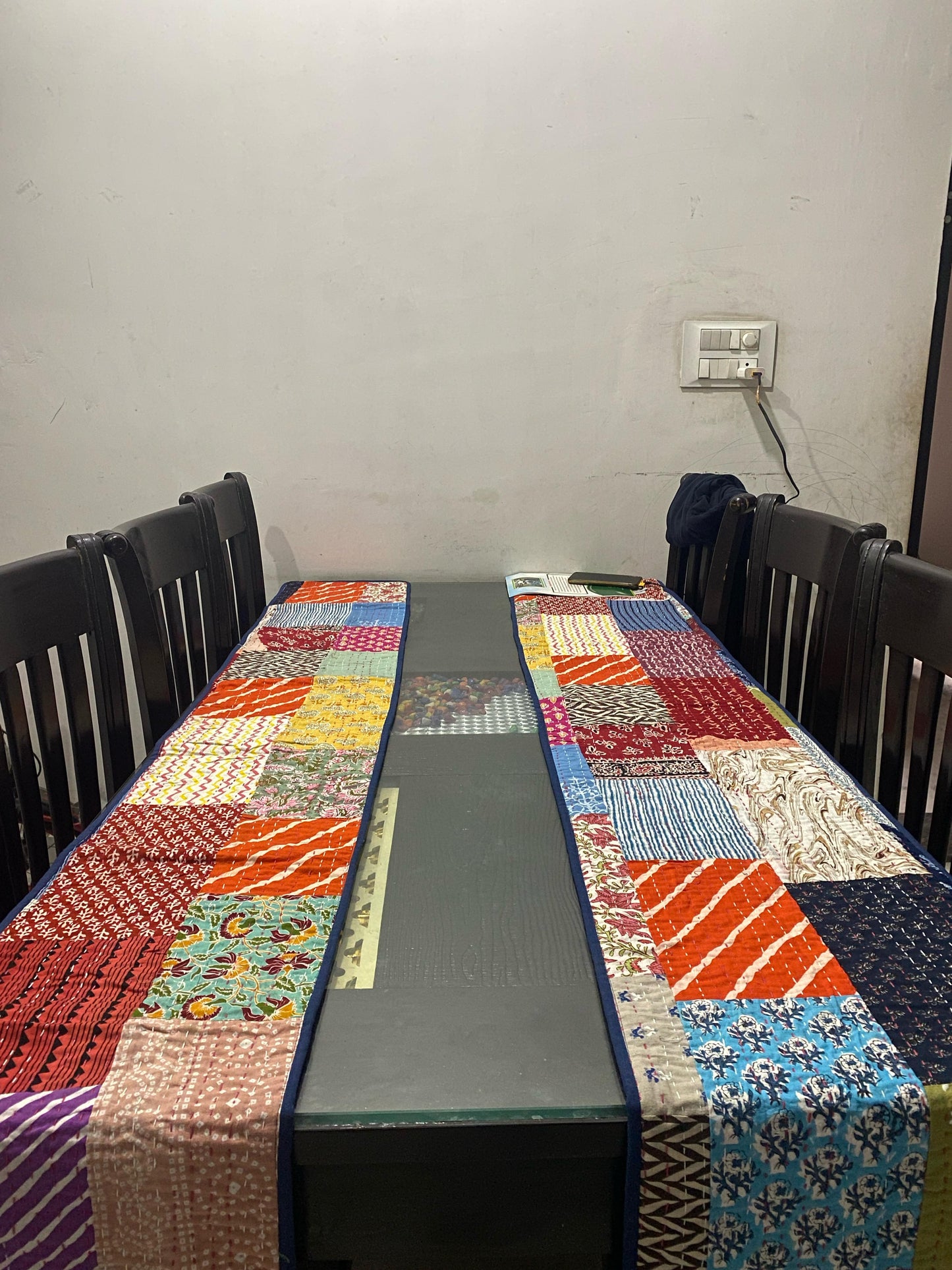 Dining table with a colorful patchwork table runner and chairs against a plain wall.