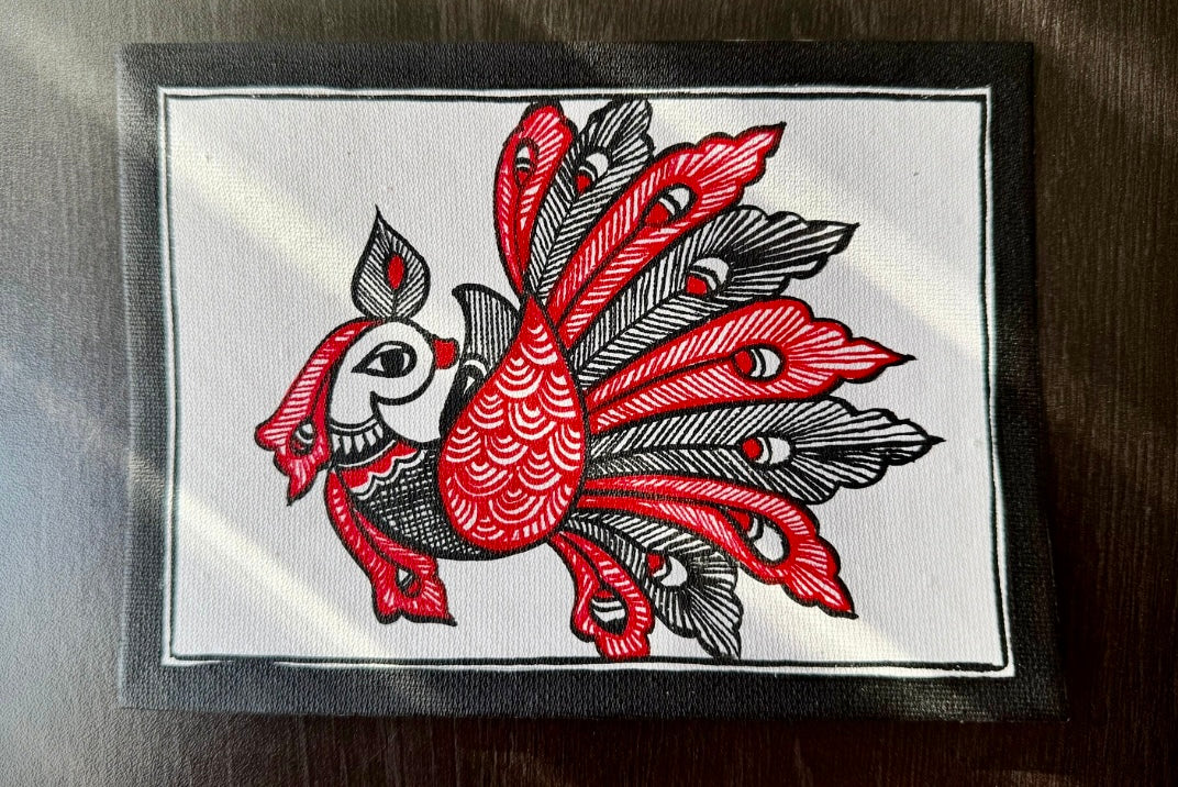 Decorative Madhubani art painting with red and black peacock on a white background, placed on a reflective surface.