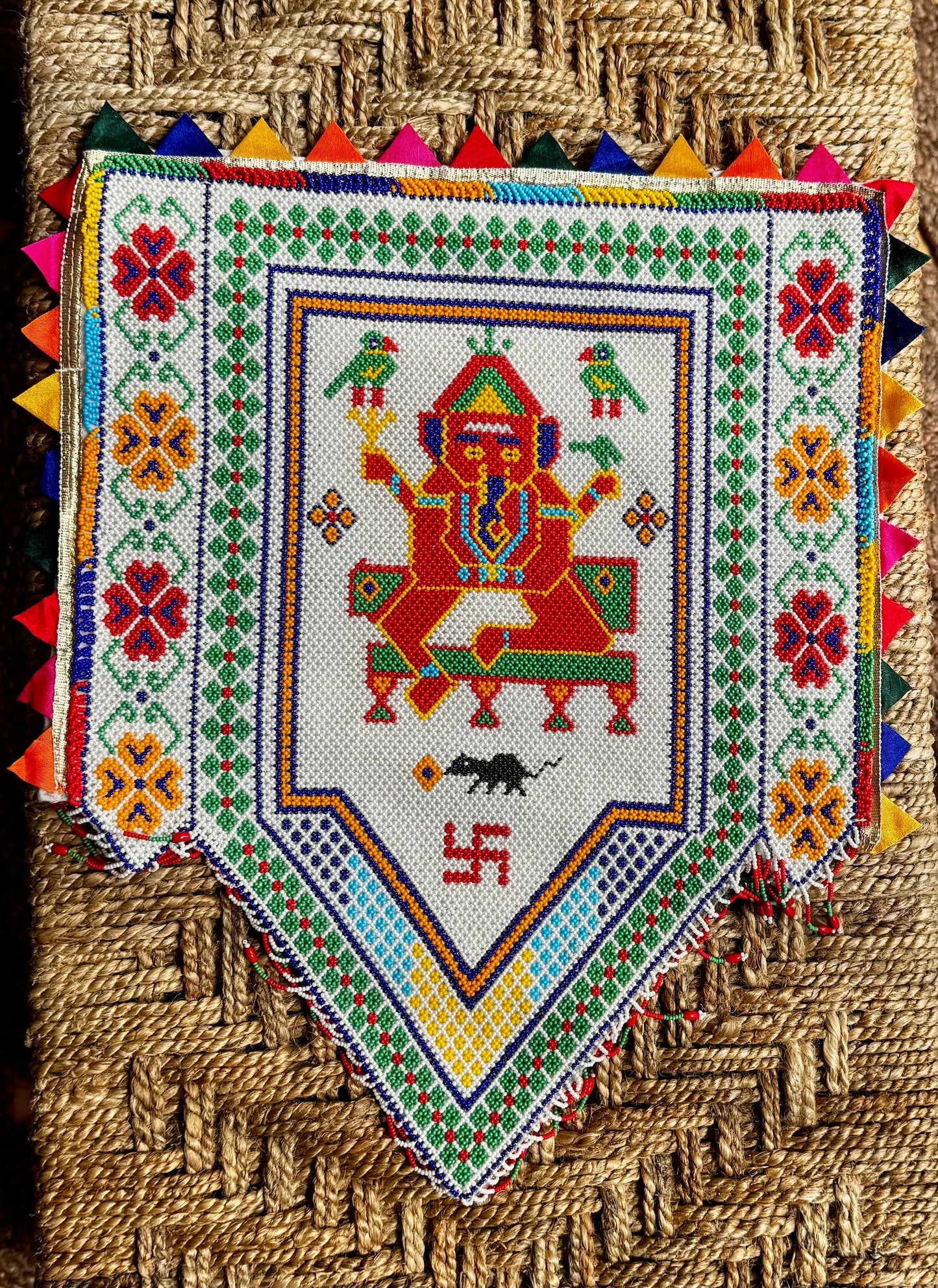 Decorative Motikaam - strung Pearl work Ganesha wall hanging with colorful design on a textured fabric background Decorative piece with a colorful design featuring a deity figure and geometric patterns on a woven background.