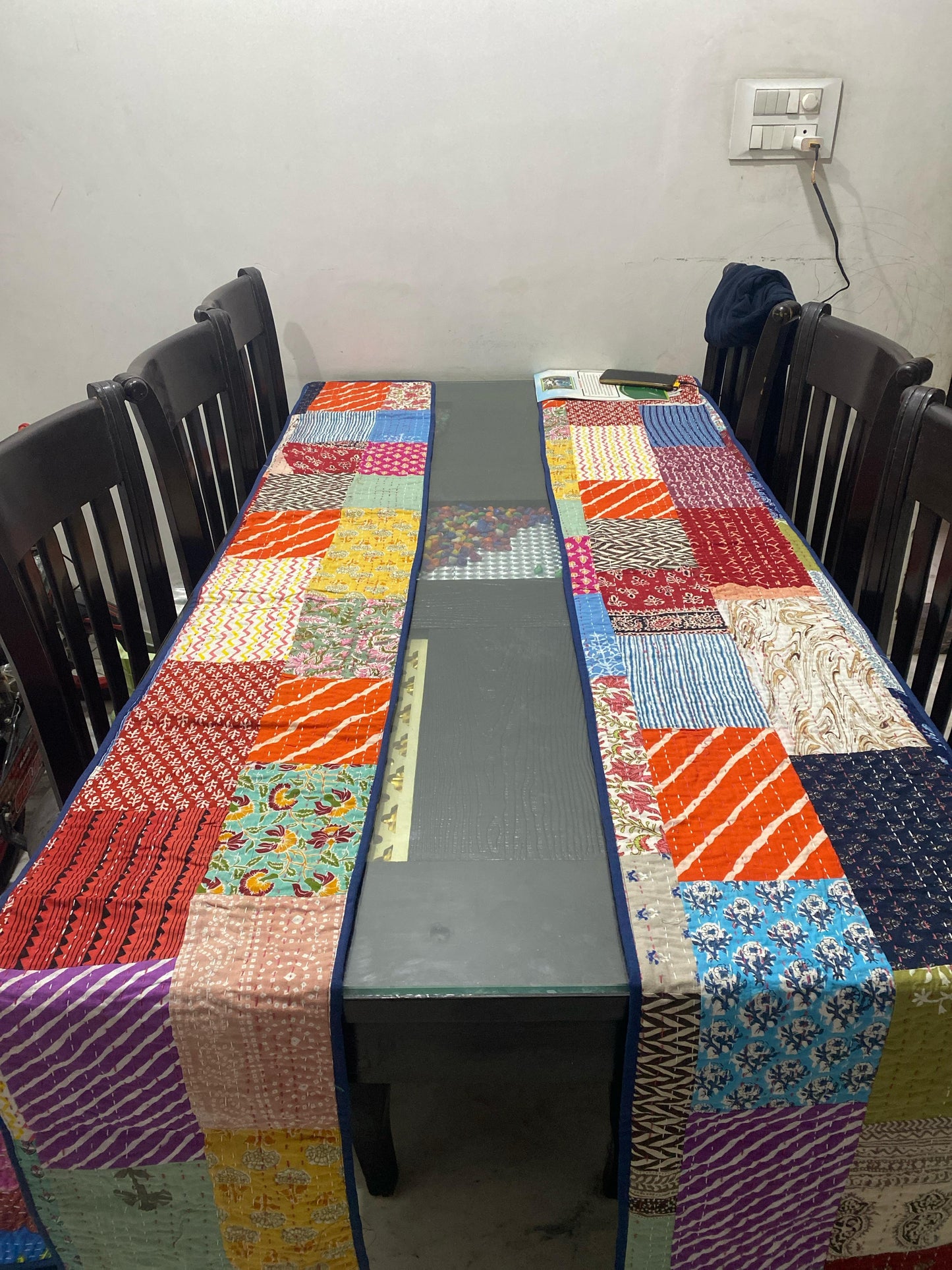 Colorful patchwork tablecloth on a dining table with dark chairs against a plain wall.