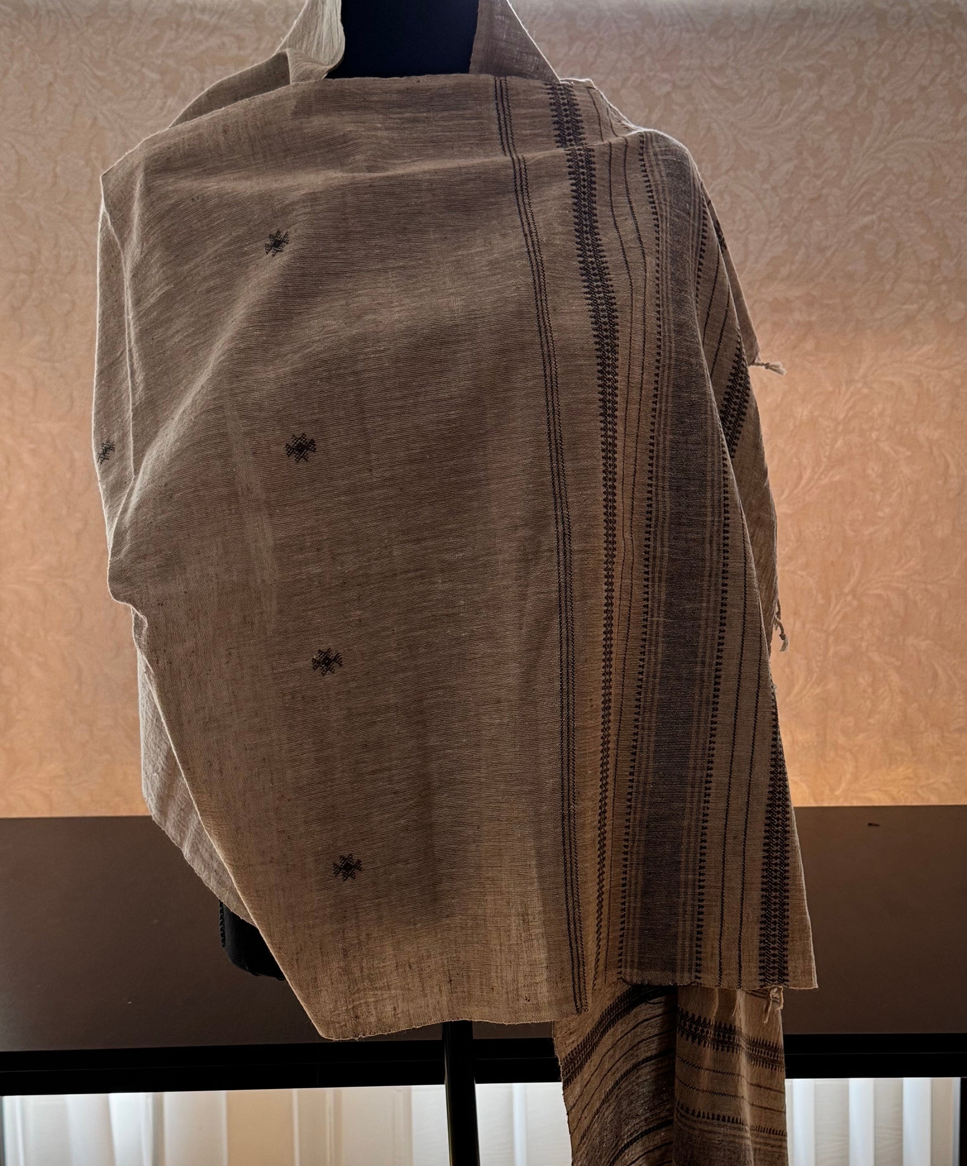 Mannequin wearing a beige cotton stole with a black weaving in a room with a window and blinds.