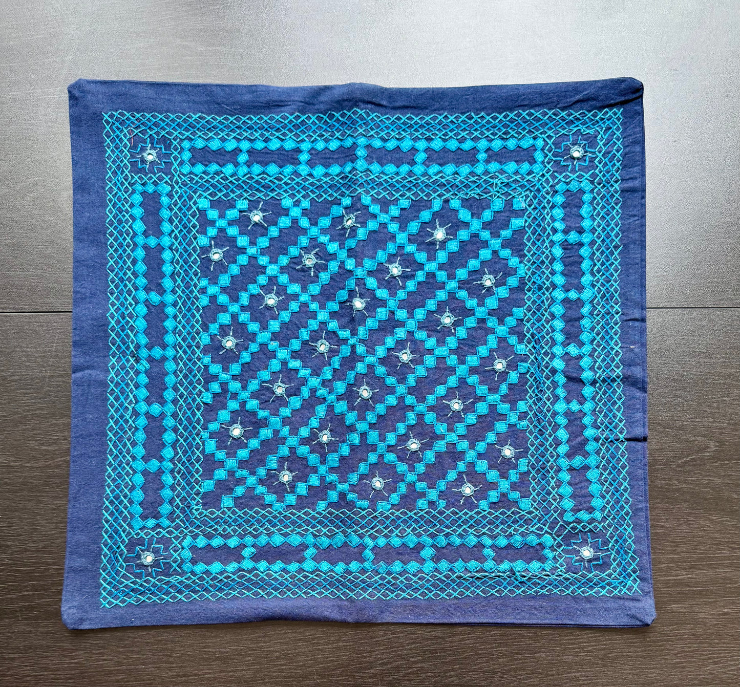 Blue cushion cover with intricate white Kutchi embroidery patterns on a wooden surface
