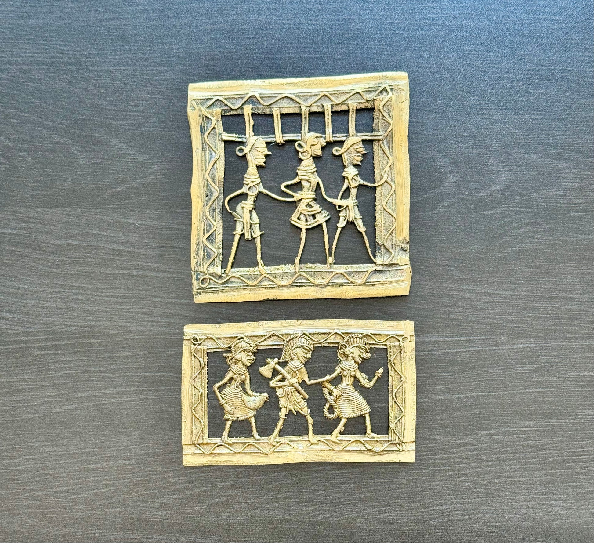 Two small square and rectangular carved brass Dhokra pieces with cutout figures, displayed on a dark gray wood-textured surface.