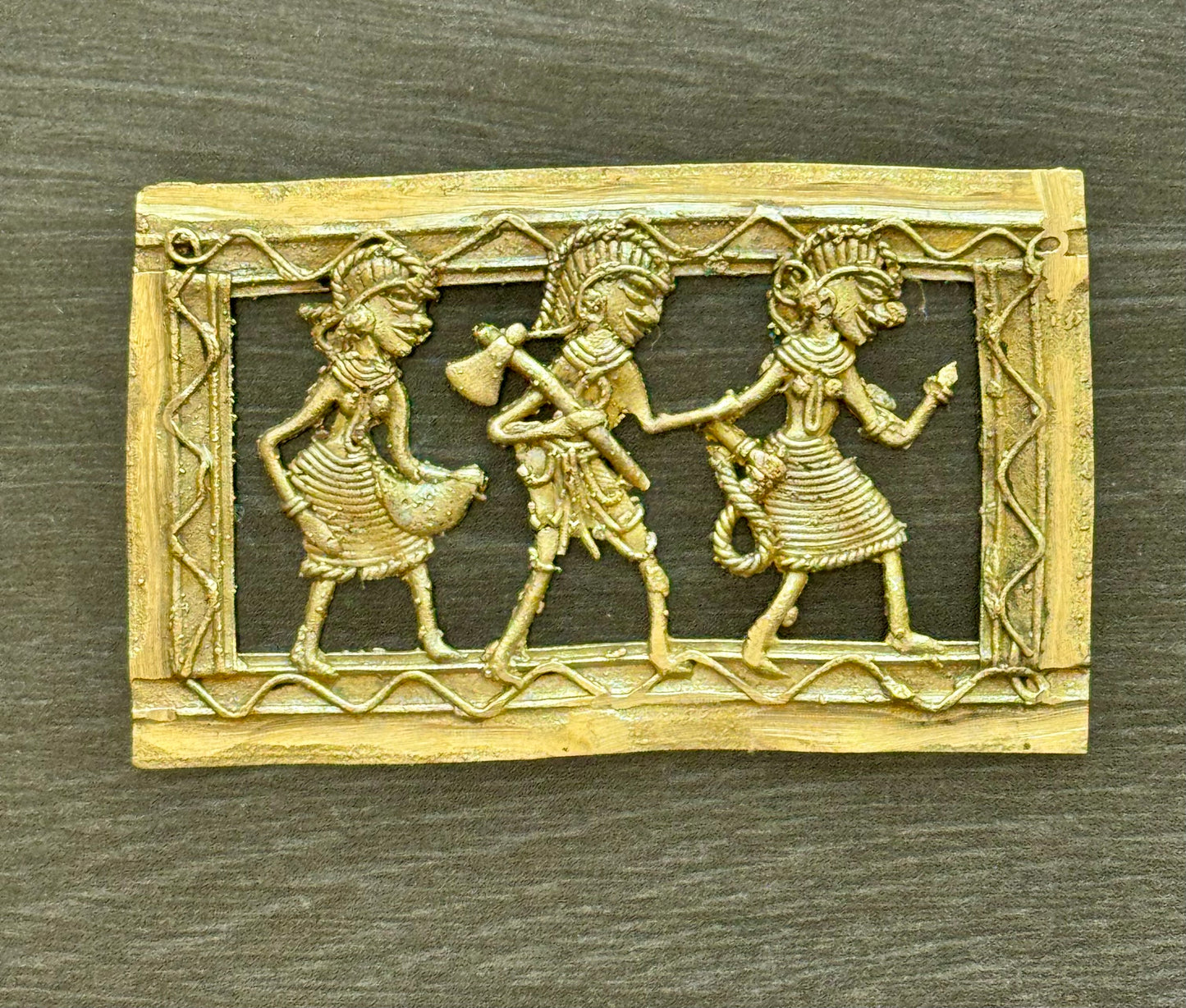 An antique-style rectangular brass Dhokra piece featuring three stylized figures in ancient  attire, set against a dark textured background.