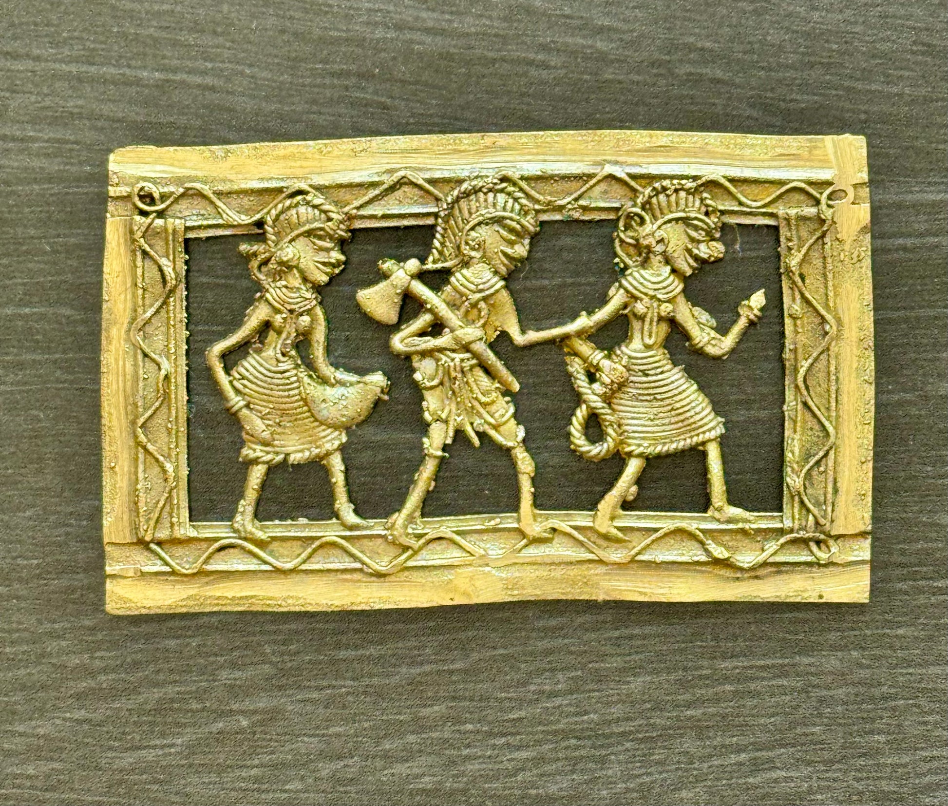 An antique-style rectangular brass Dhokra piece featuring three stylized figures in ancient  attire, set against a dark textured background.