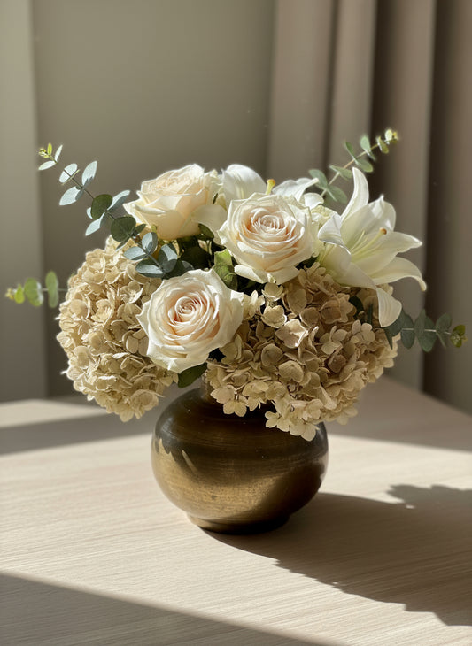 Small brown brass pot vase on a dark surface with light filtering through blinds in the background