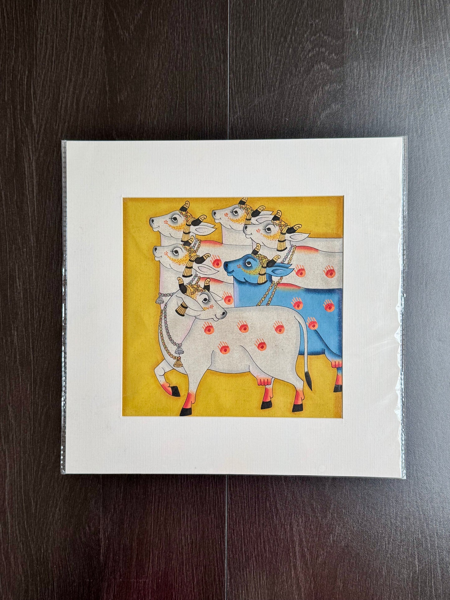 Framed Pichwai artwork of a colorful depiction of cows on a yellow background.