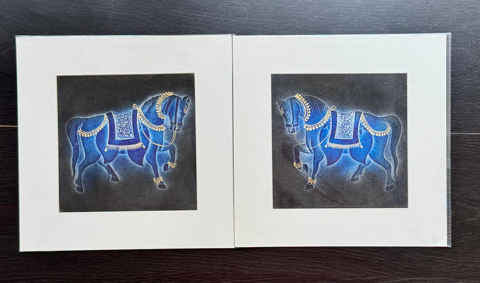 Two nail drawn paintings of blue horses with gold detailing on a black background, framed together.