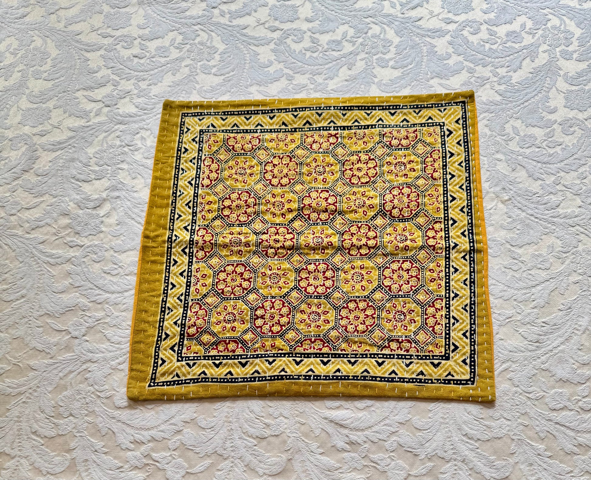 Decorative yellow and red patterned pillow in Ajrakh hand block print fabric on a textured white background