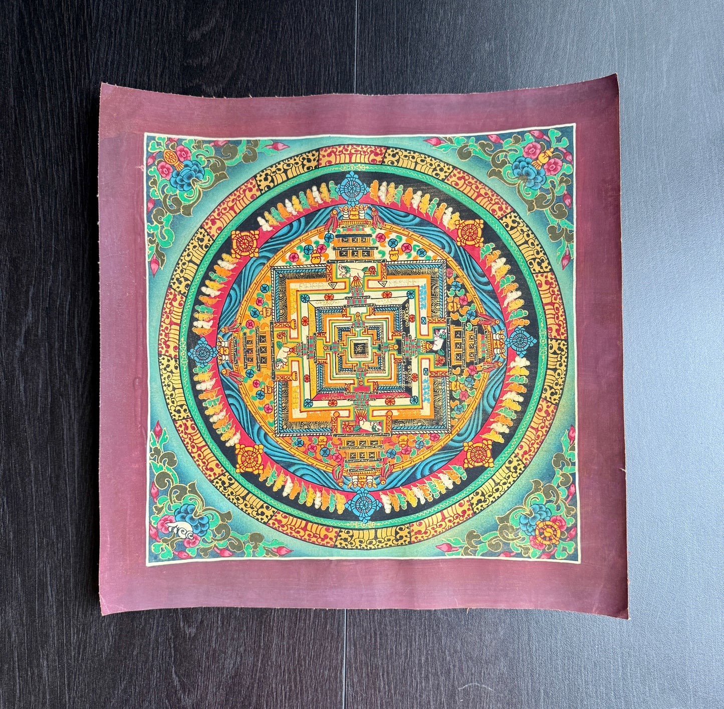 Colorful mandala design in Thangka art style on a square against a dark wooden surface.