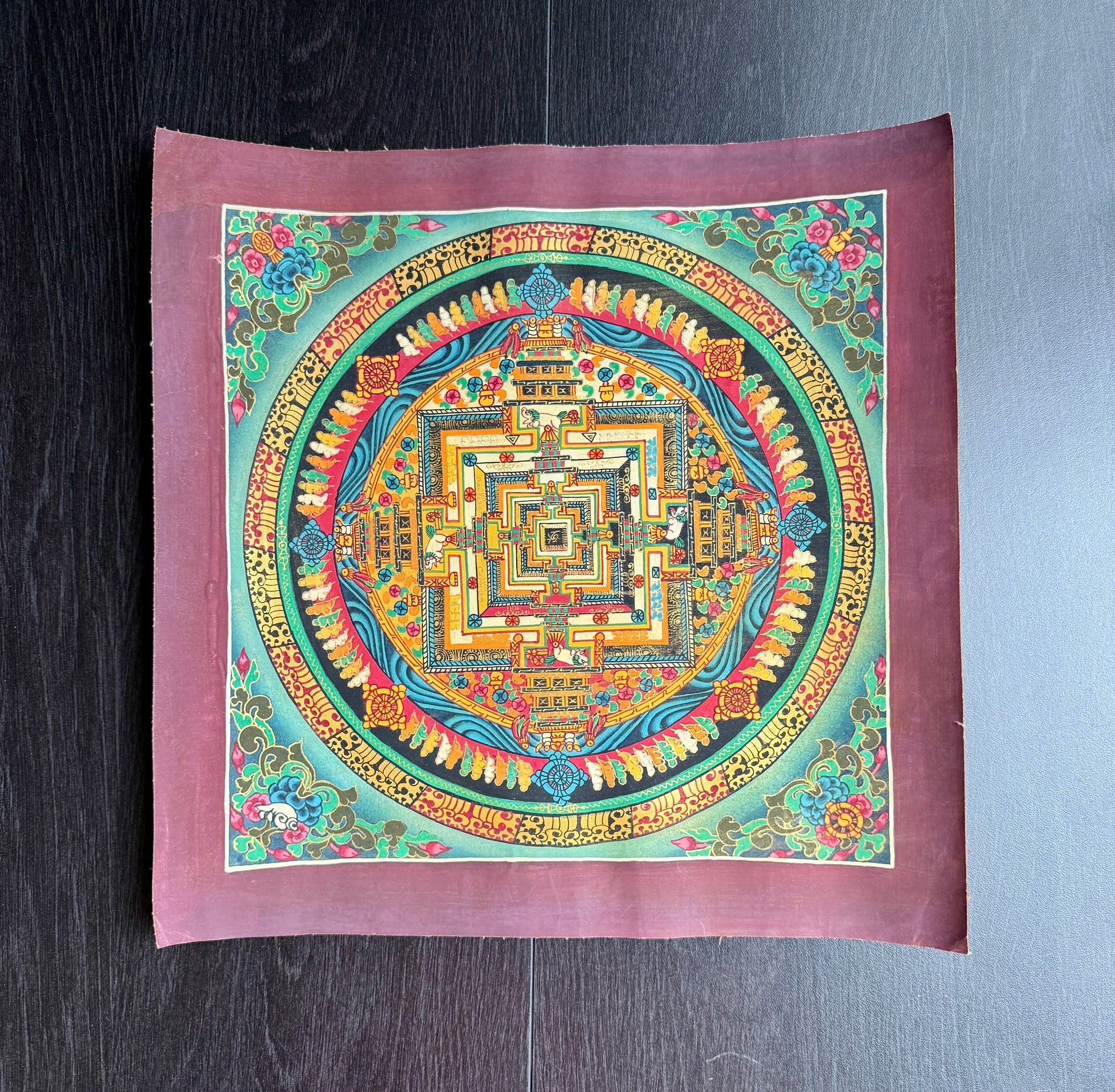 Colorful mandala design in Thangka art style on a square against a dark wooden surface.