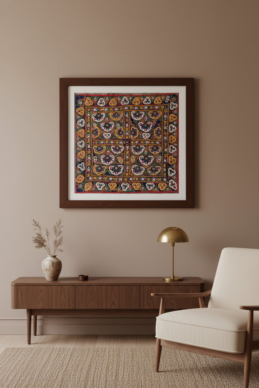 Framed Kutchi textile tapestry on a wall above a wooden console table with a chair and lamp.