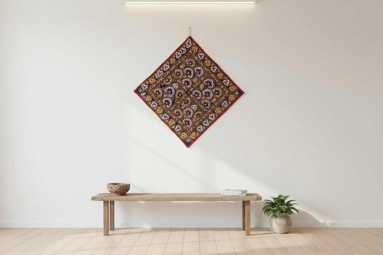 Decorative Kutchi tapestry with floral pattern hanging on a white wall above a wooden bench.