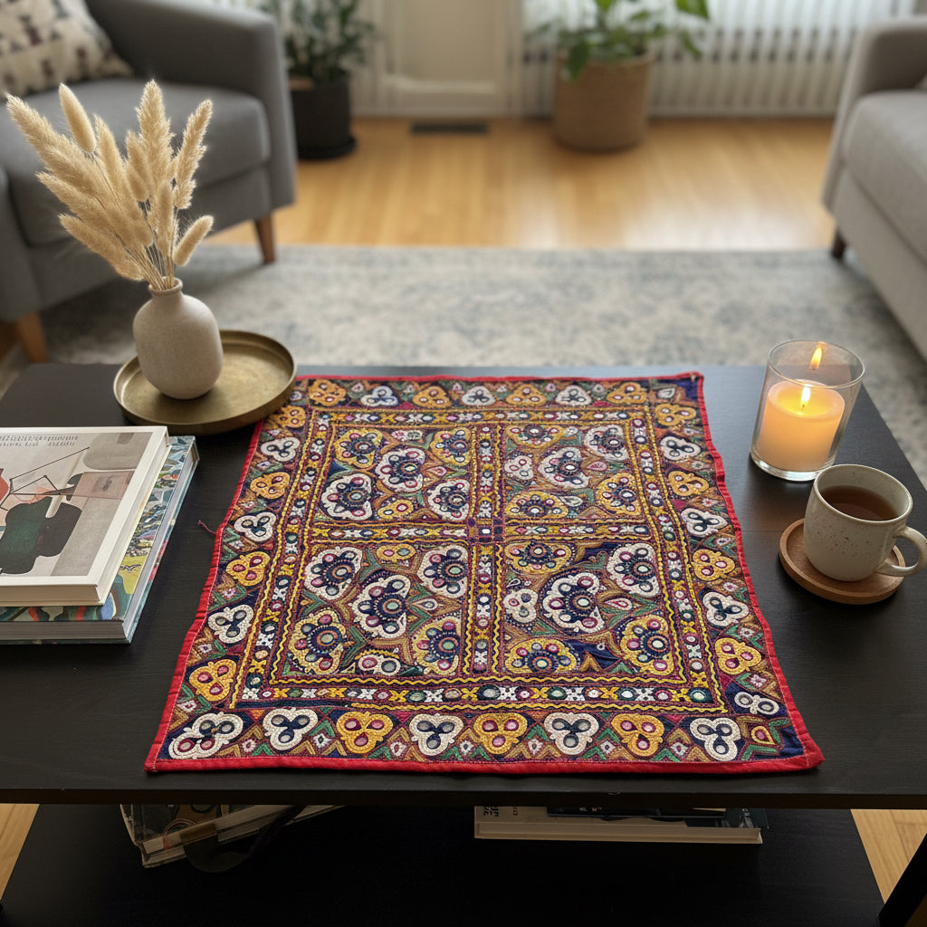 Decorative table setting with a colorful Kutchi embroidered Textlle, books, a cup, and candle on a dark surface.