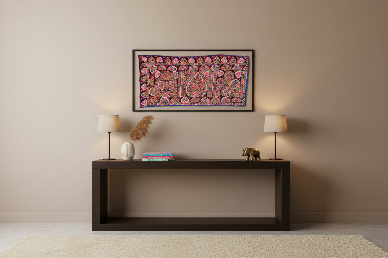 Decorative Kutchi textile wall art above a wooden console table with lamps and books in a room.