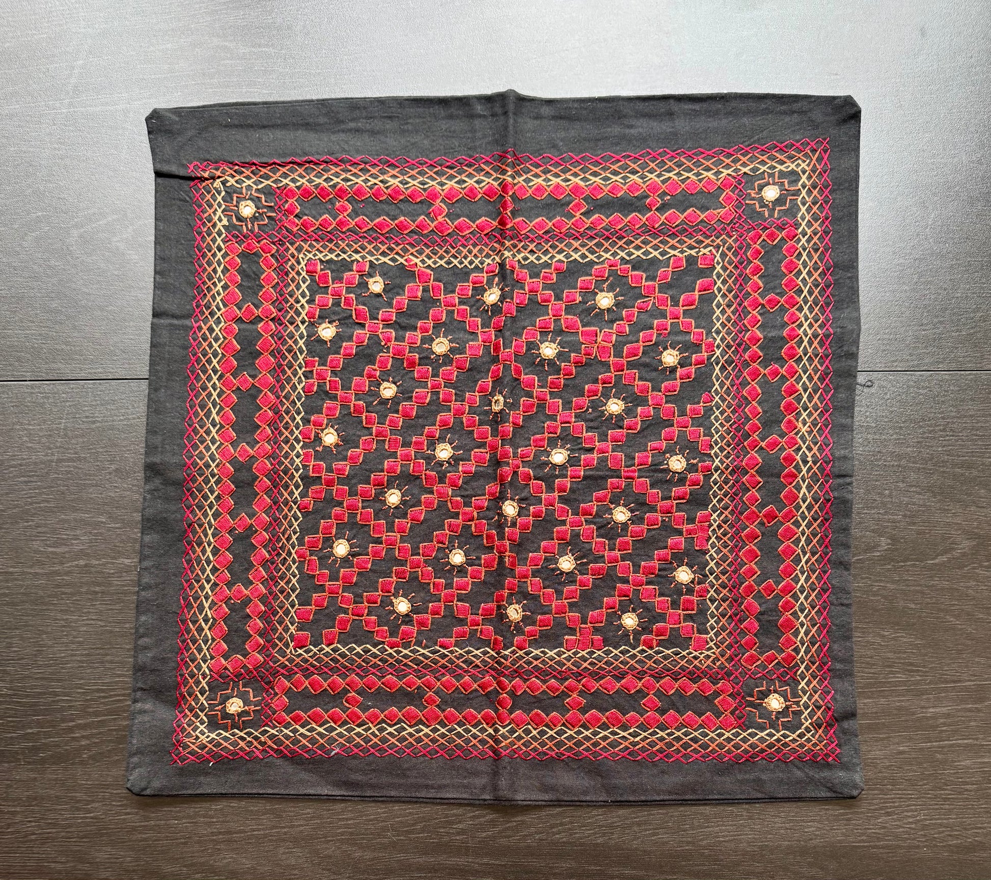 Decorative fabric cushion cover with red and black Kutchi embroidery pattern on a wooden surface