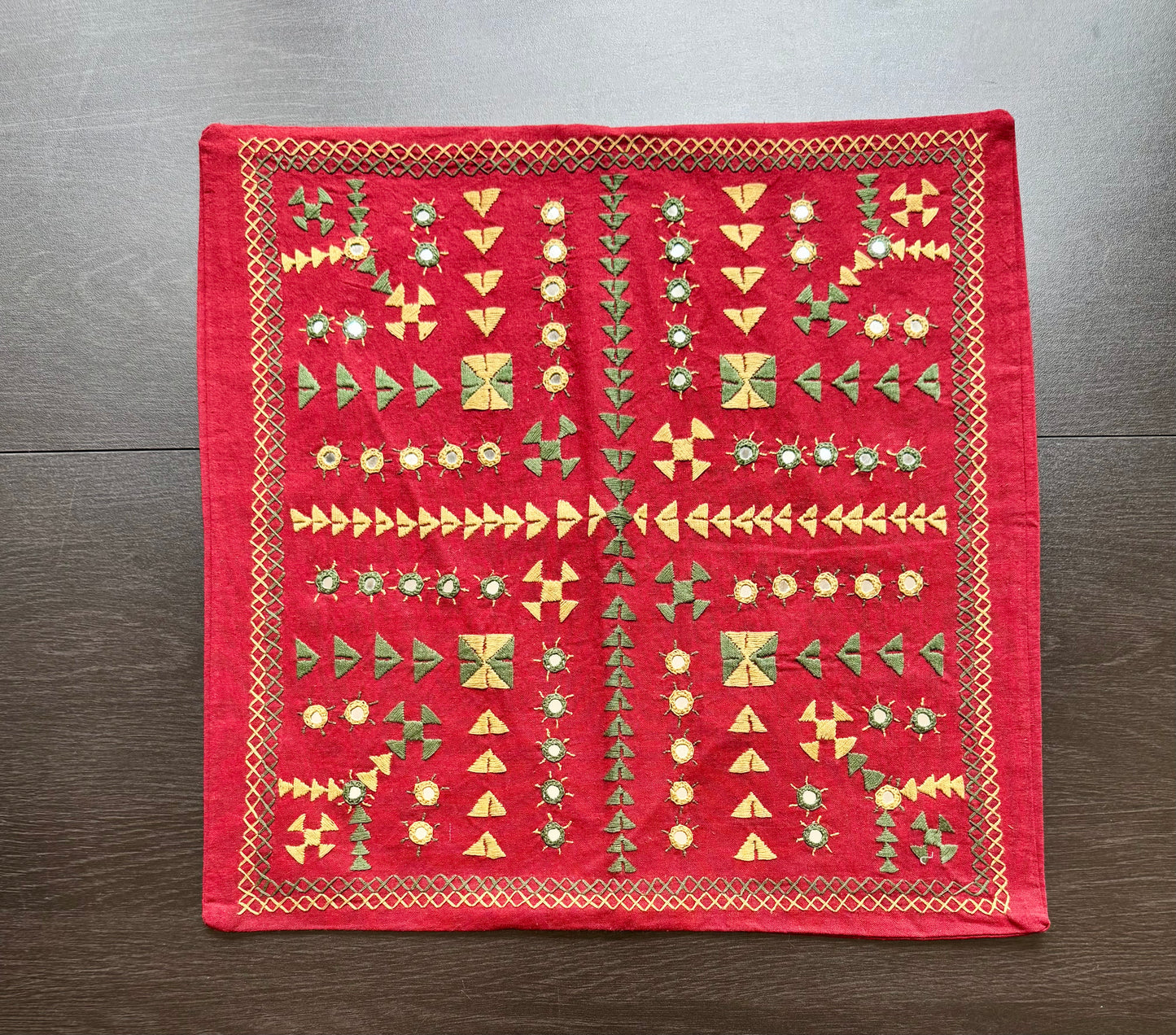 Red embroidered fabric cushion cover with geometric Kutchi embroidery patterns on a wooden surface