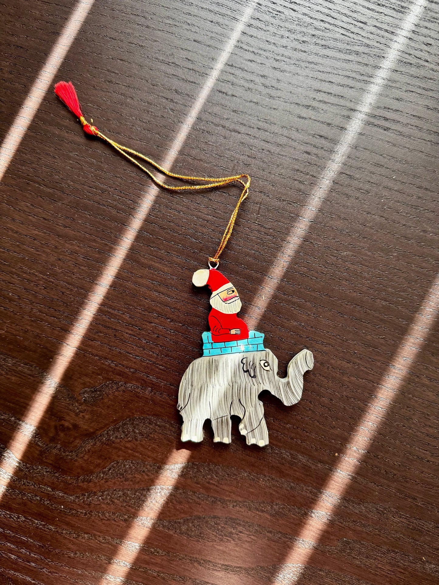 Decorative ornament with a Santa Claus figure on an elephant, hanging from a string on a wooden surface.