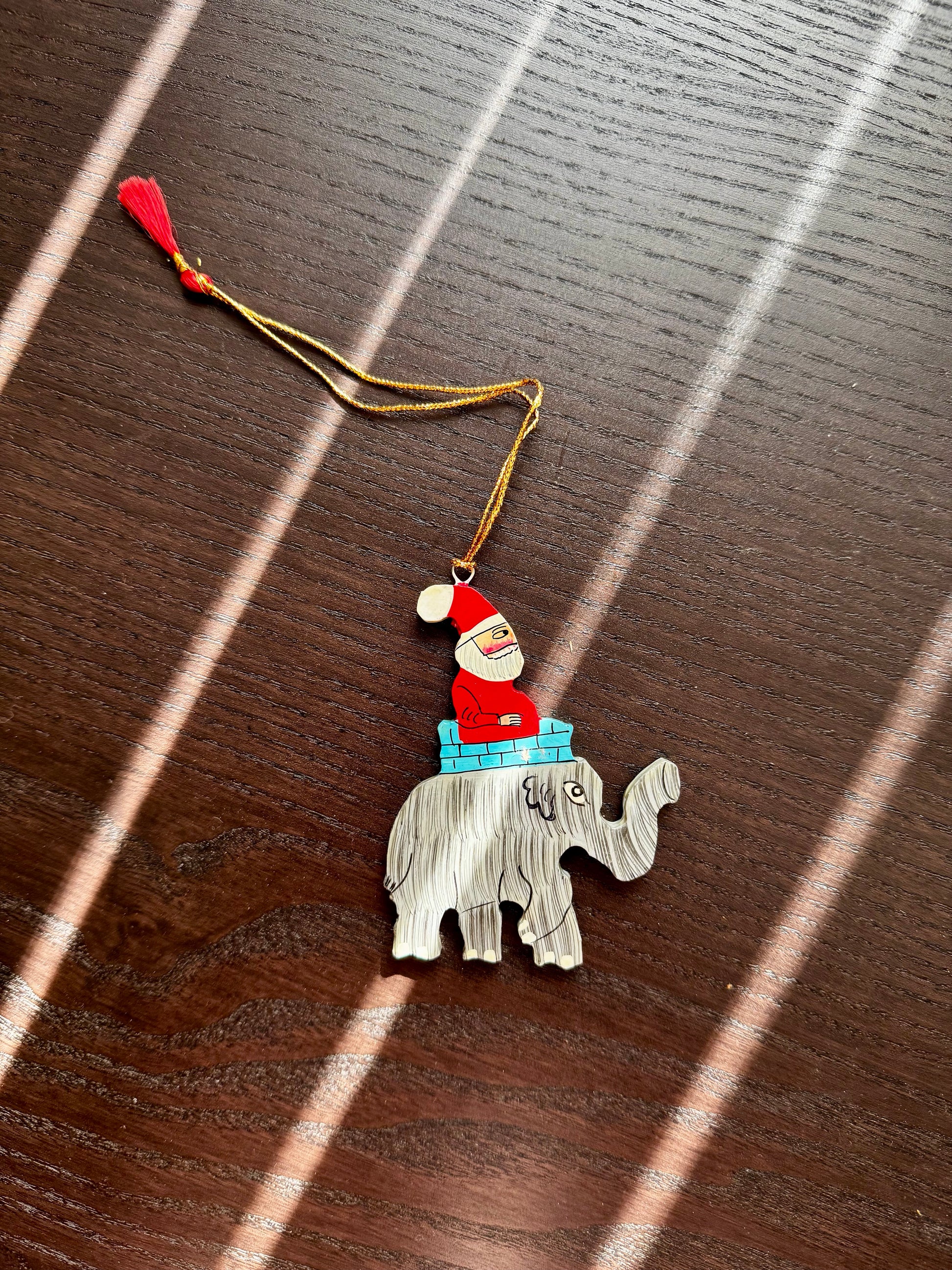 Decorative ornament with a Santa Claus figure on an elephant, hanging from a string on a wooden surface.