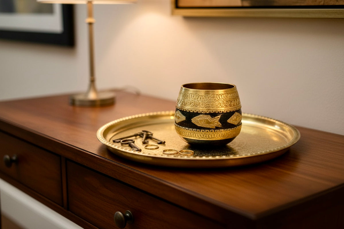 A small ornate brass-toned Sherpai bowl with black decorative bands sits on a matching round gold tray placed on a wooden dresser in a warmly lit room.