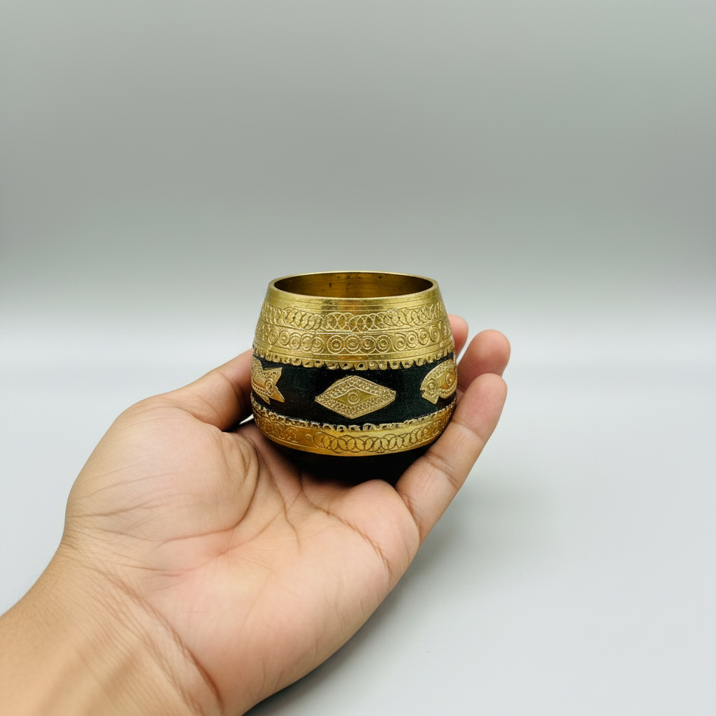 A hand holding a small decorative wood and brass Sherpai small bowl with a black band and intricate gold geometric patterns, set against a neutral gray background.