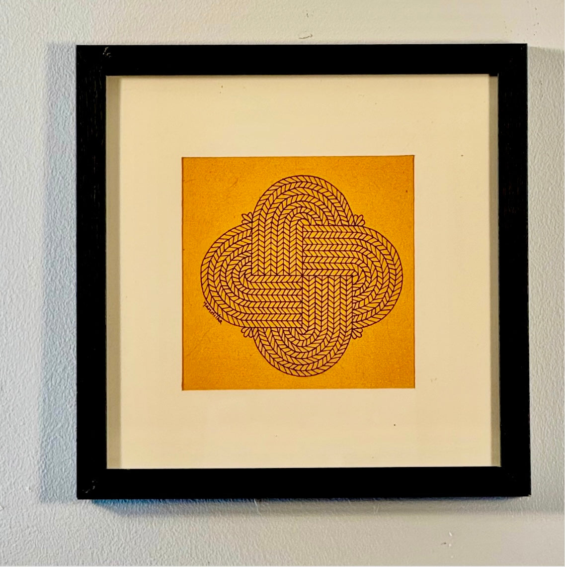 Framed Chittara art featuring a central interlocking geometric knot design in brown on a mustard yellow square background, surrounded by a wide cream mat and a black frame against a light gray wall.