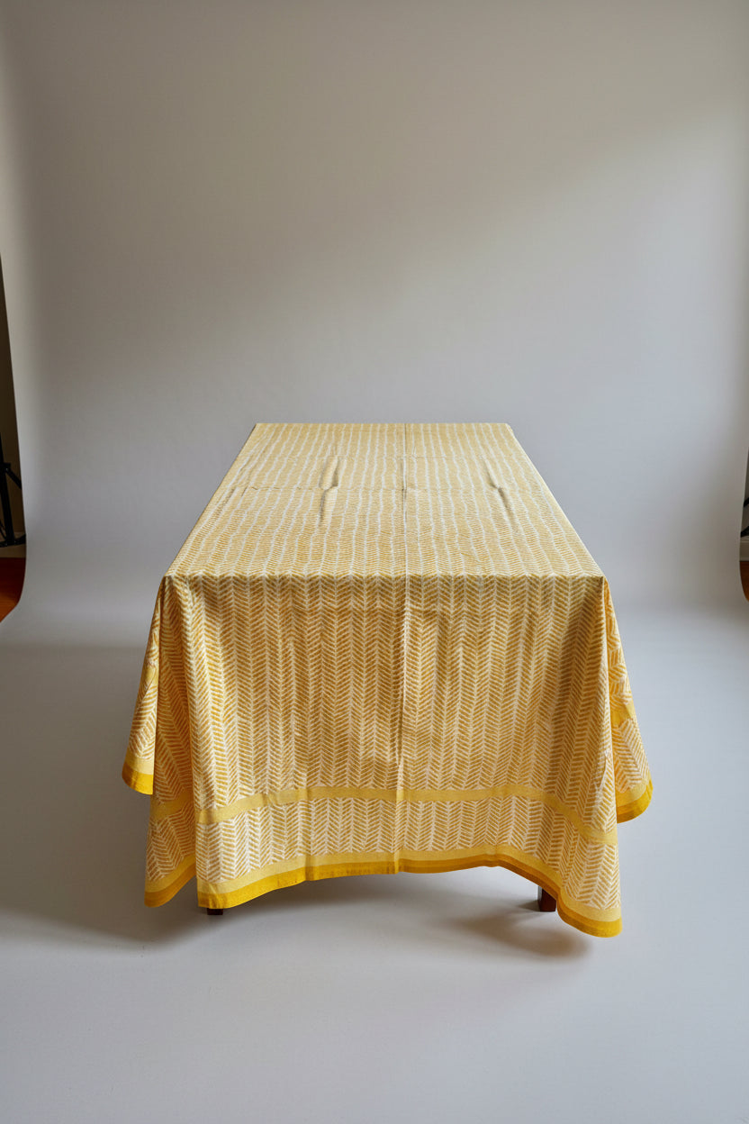 Living room with a table covered by a yellow hand block print tablecloth, wooden floor, and curtains.